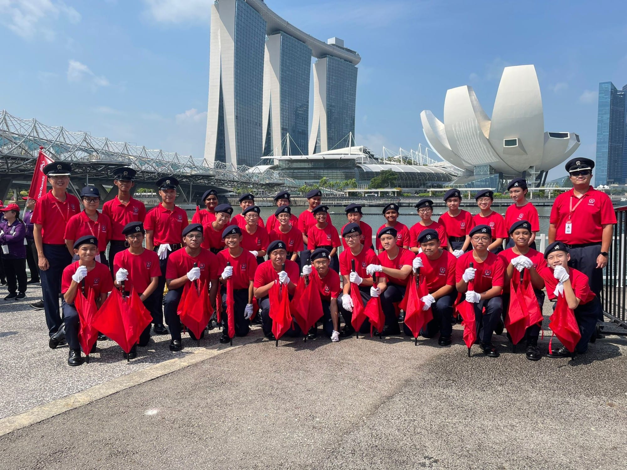 A large group in red uniforms and black berets holding red umbrellas pose in front of Marina Bay Sands in Singapore.
